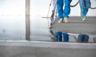 Ground level of faceless site constructors in overall holding hose while pouring liquid self leveling concrete on floor in building